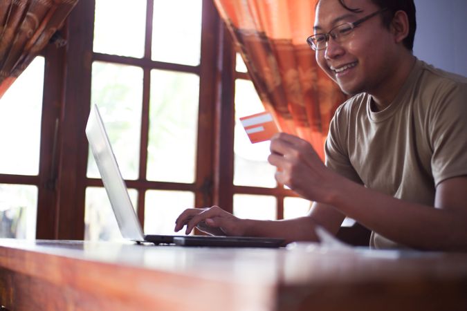 Asian Male with shirt doing online transaction in living room with credit card smartphone and laptop