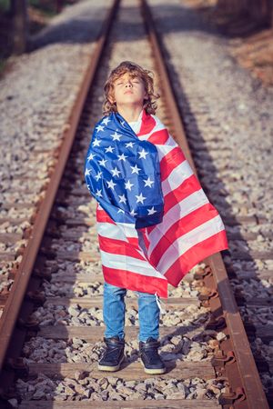 Adorable preteen boy wrapped in American flag standing on railwa