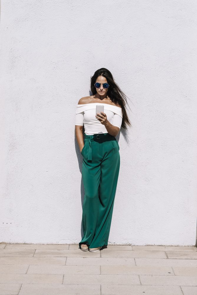 long haired woman with sunglasses leaning on a white wall while using a mobile phone in a sunny day