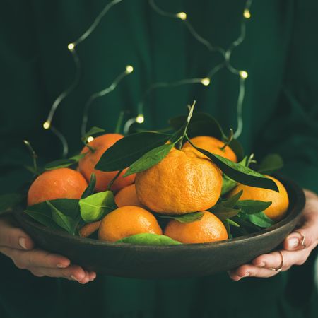 Woman in green holding bowl of tangerines, with fairy lights, square crop