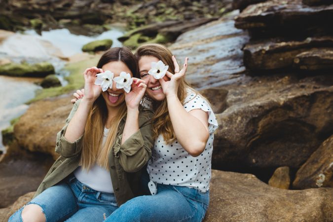 Young female travelers sitting on stone and covering eyes with flowers