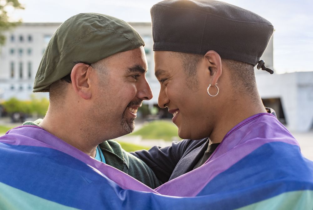 Gay couple under LGBT rainbow flag while laughing and looking at each other. Concept of Pride. Pride month.