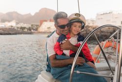 Man hugging smiling child and sitting near steering wheel on yacht in sea