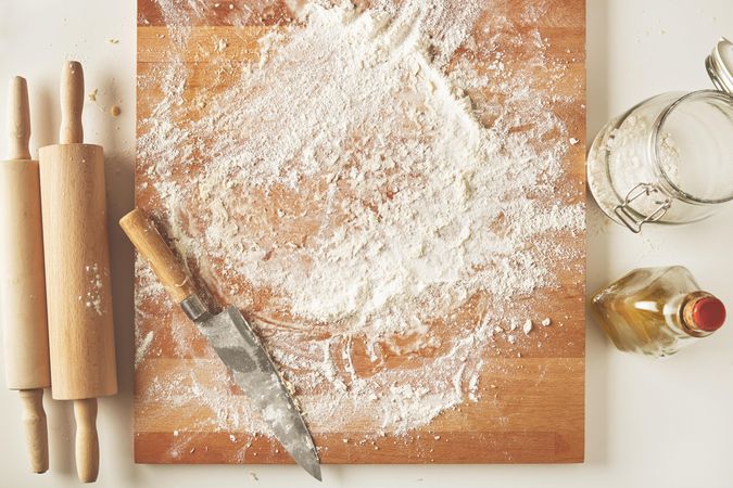 Breadboard scattered with flour, a knife, and rolling pins and olive oil to the side