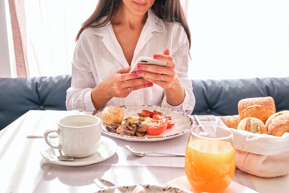 Crop female at table with served breakfast and browsing smartpho