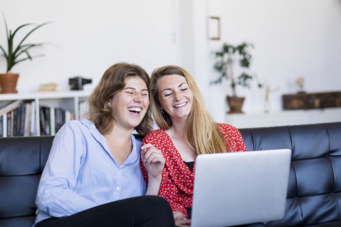 Two young women using computer while sitting on couch in living room
