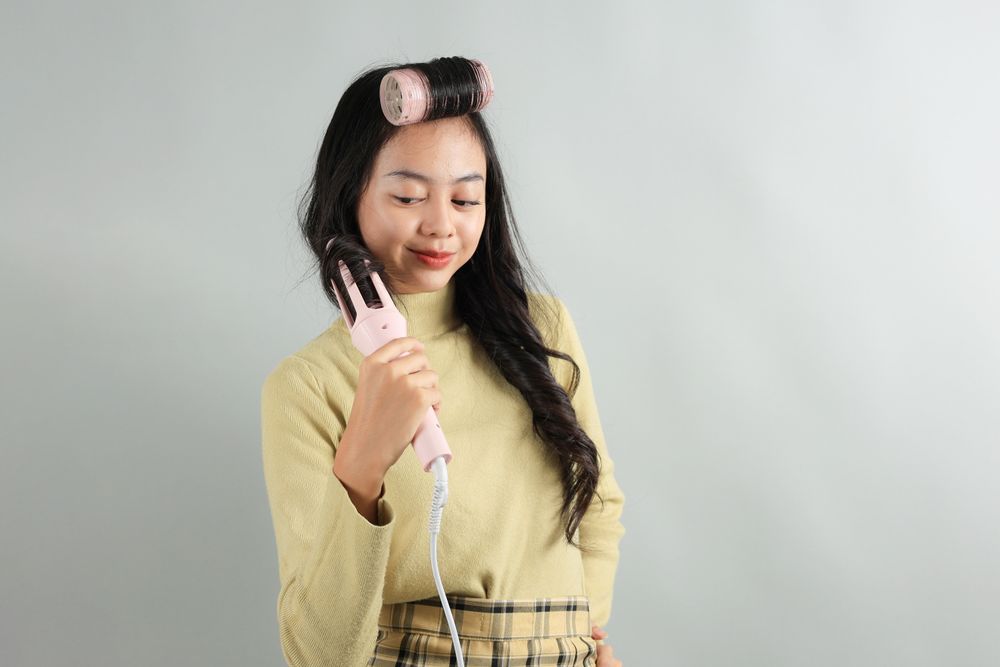 Asian Teenager using Curling Hair Iron on Grey Background.