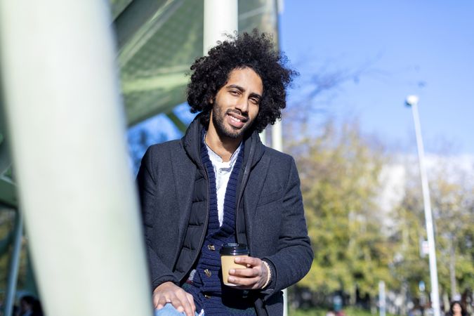 Content Black man in jacket holding warm beverage sitting outside