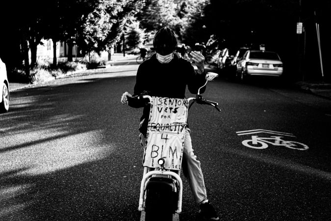 Grayscale photo of person with facemask riding a motorcycle with banner for Black Lives Matter protest