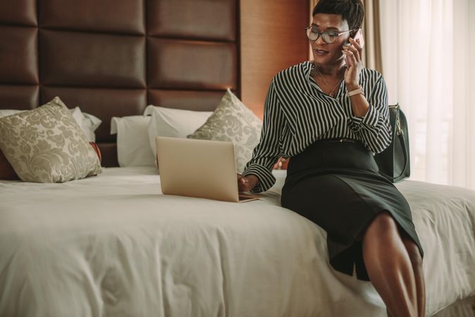 Businesswoman in hotel room using laptop and smartphone sitting on bed