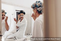 Two women in bathrobes applying cream on face standing in front of mirror 0KPPM5