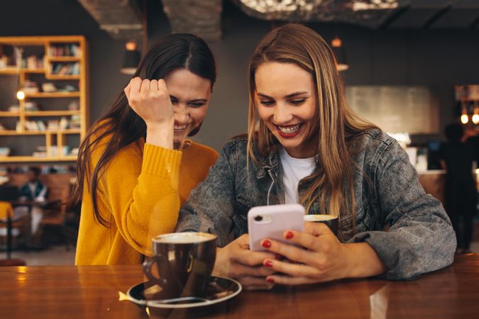 Happy female friends at a cafe using smart phone with a cup of coffee on the table