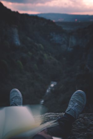 View to small river in canyon and legs of person with book sitting on the edge