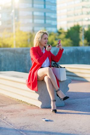 Trendy blonde woman applying lipstick sitting on a bench and using the smartphone as a mirror