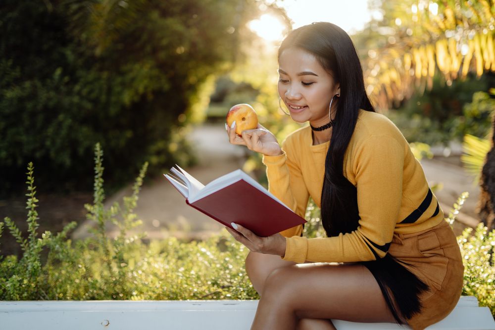 Graceful long haired Asian woman eating apple and reading book