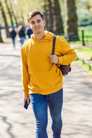 Smiling man in yellow hoodie with backpack walking in park, vertical