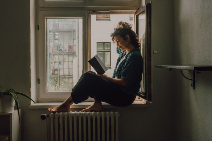 Interested woman reading book at window sill at home