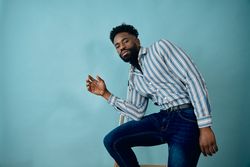 Black man in striped blue shirt and jeans in blue studio leaning on step ladder