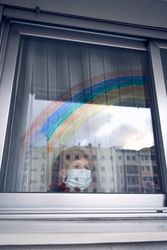 Boy in mask standing near window with rainbow at home
