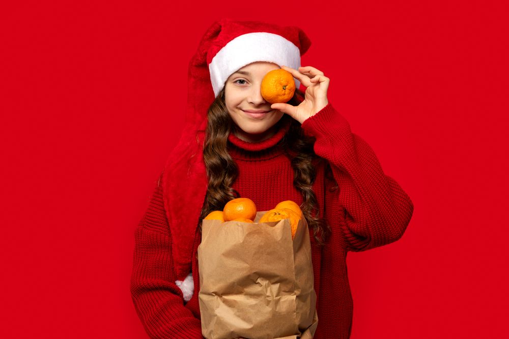 Girl in Santa hat holds orange in front of eye.