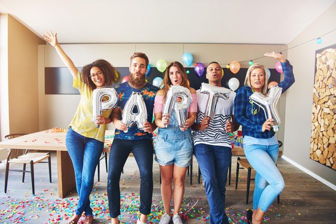 Friends standing in room with confetti fallen on floor and silver “party” balloons