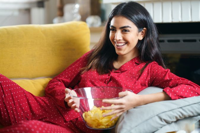 Female in red pajamas lying on sofa and eating chips