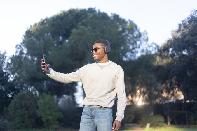 Black male with sunglasses standing in park taking selfie with phone
