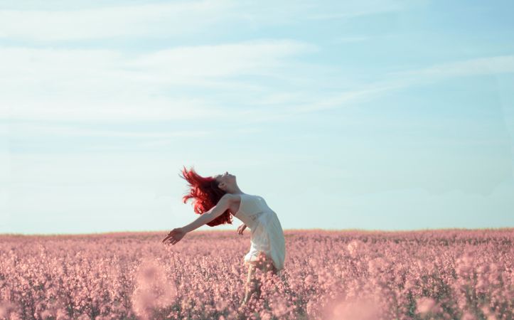 Side view of woman with red hair wearing light dress and bending on pink flower meadow