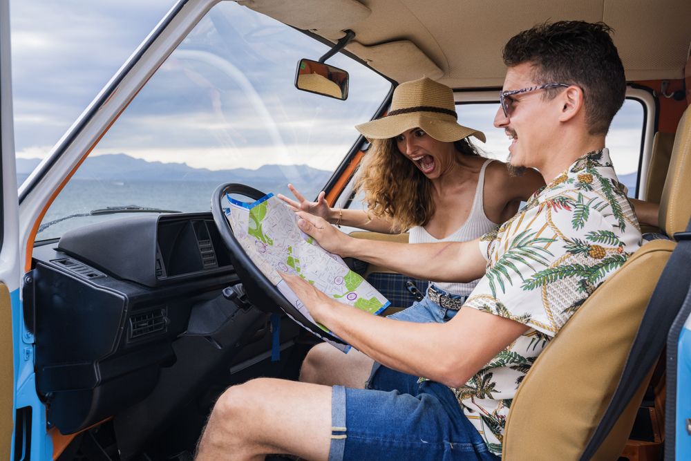 Touristic man exploring map with help of long haired woman in hat in front seat of car