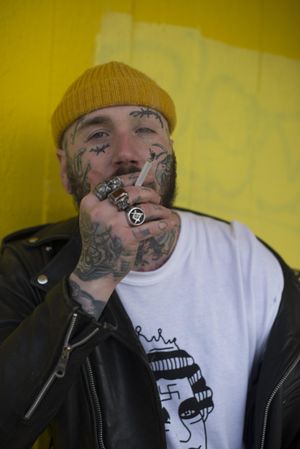 Punk hipster man wearing beanie with multiple facial tattoos smoking cigarette against yellow wall