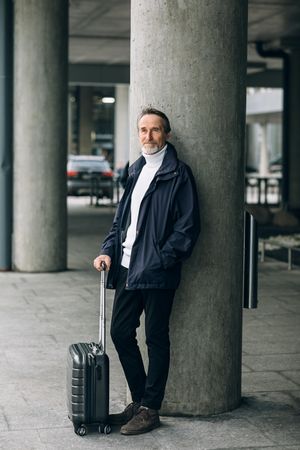 Mature man with suitcase leaning against concrete pillar, vertical