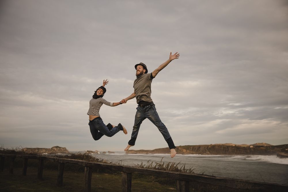Couple holding hands and jumping on bench on shore near water
