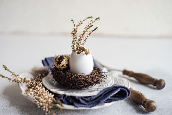 Side view of Easter table setting with decorative egg shell, feather and heather on plates on grey counter with copy space