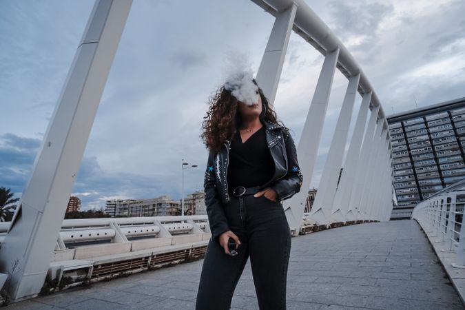 Stylish woman in jacket and jeans smoking on bridge