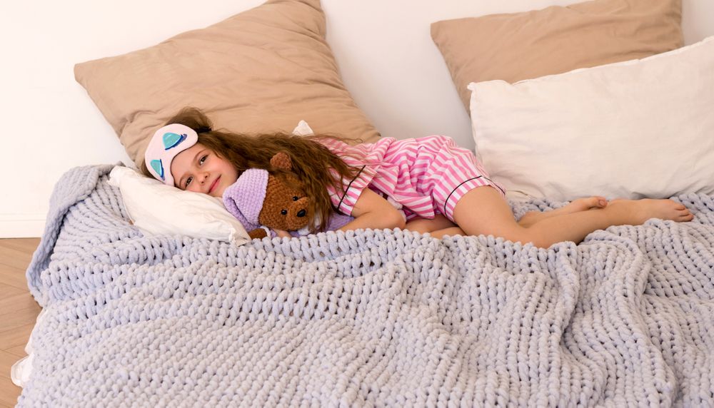 Girl in sleep mask with teddy bear lying on bed.