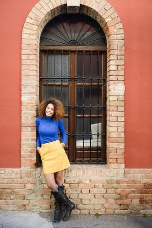 Female with curly hair wearing bright blue shirt and yellow skirt standing in front of large window