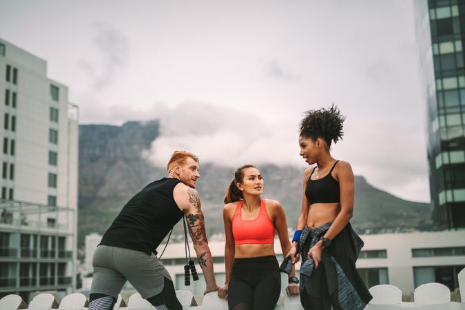 Two fitness women with a man relaxing after workout on rooftop and talking to each other