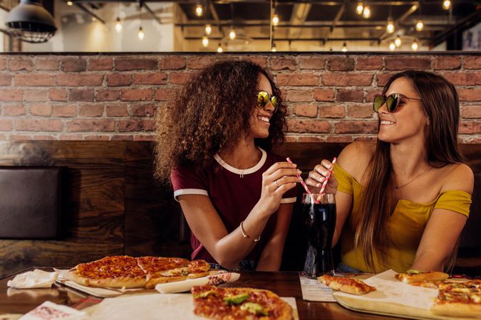 Two females drinking cold drink at cafe with pizza on table