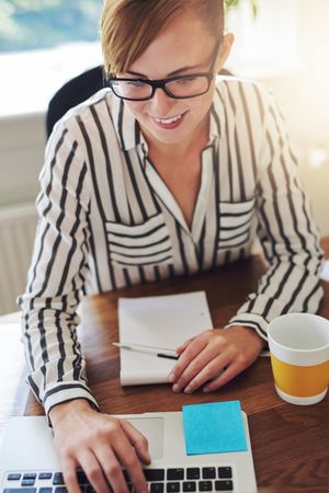 Content female in striped shirt with coffee working on laptop at her desk