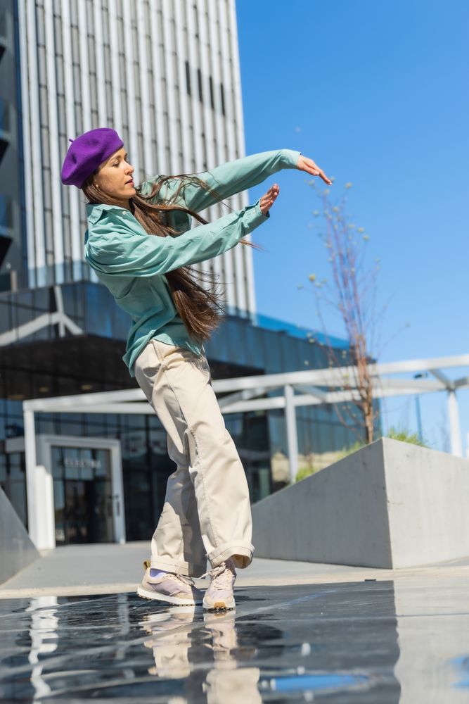 A Young Woman In A Purple Beret And Teal Jacket Performs A Dynamic Dance Move On A Wet Urban Plaza, Reflecting The Sky And Buildings Around Her.