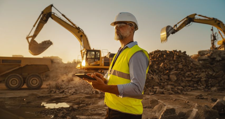 Male Civil Engineer Wearing Protective Goggles And Using Tablet On Construction Site On Sunny Day. Man Inspecting Building Progress. Excavator Loading Materials Into Big Industrial Truck.