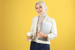 Smiling Young Asian Hijab Woman in Cream Suit and Hijab Holding a Laptop and Hot Coffee Cup.