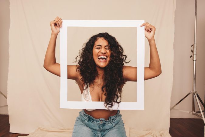 Happy young woman laughing through a white picture frame in a studio