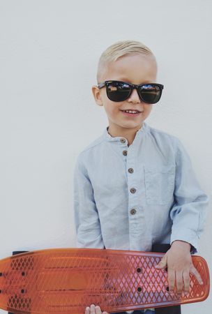 Young trendy blond boy leaning against a wall with red skateboard, vertical