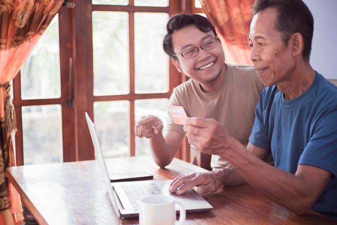 Happy adult son and older father pay online using laptop and credit card
