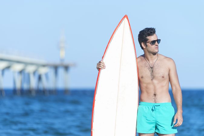 Male surfer standing with red outlined board on coastline