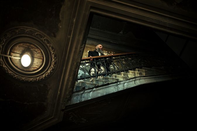 Bearded man in dark jacket standing on staircase in an old building