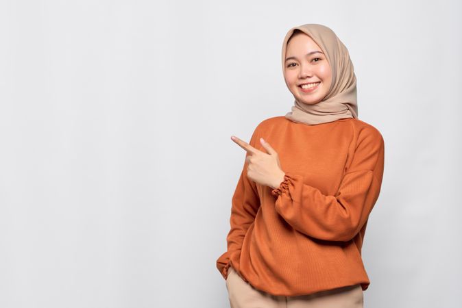 Muslim woman in headscarf and orange sweater pointing to copy space on her side while smiling
