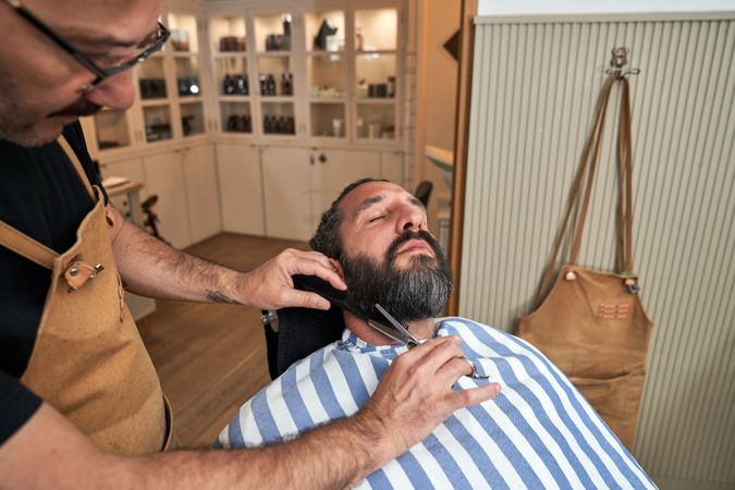 Barber cutting beard of client in barbershop