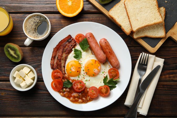 Wooden table with breakfast plate of coffee, eggs, tomatoes, sausage and bacon with cutlery